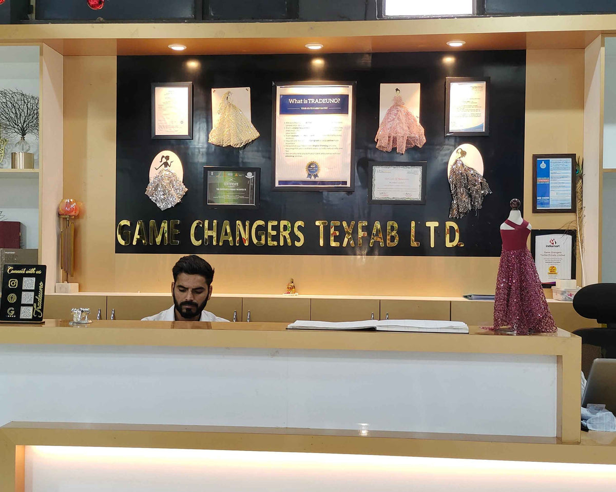 A modern textile factory reception desk with a man working behind it. The desk is white with light wood trim, and the back wall features a black panel with gold lettering that reads 'GAME CHANGERS TEXTILE LTD'.