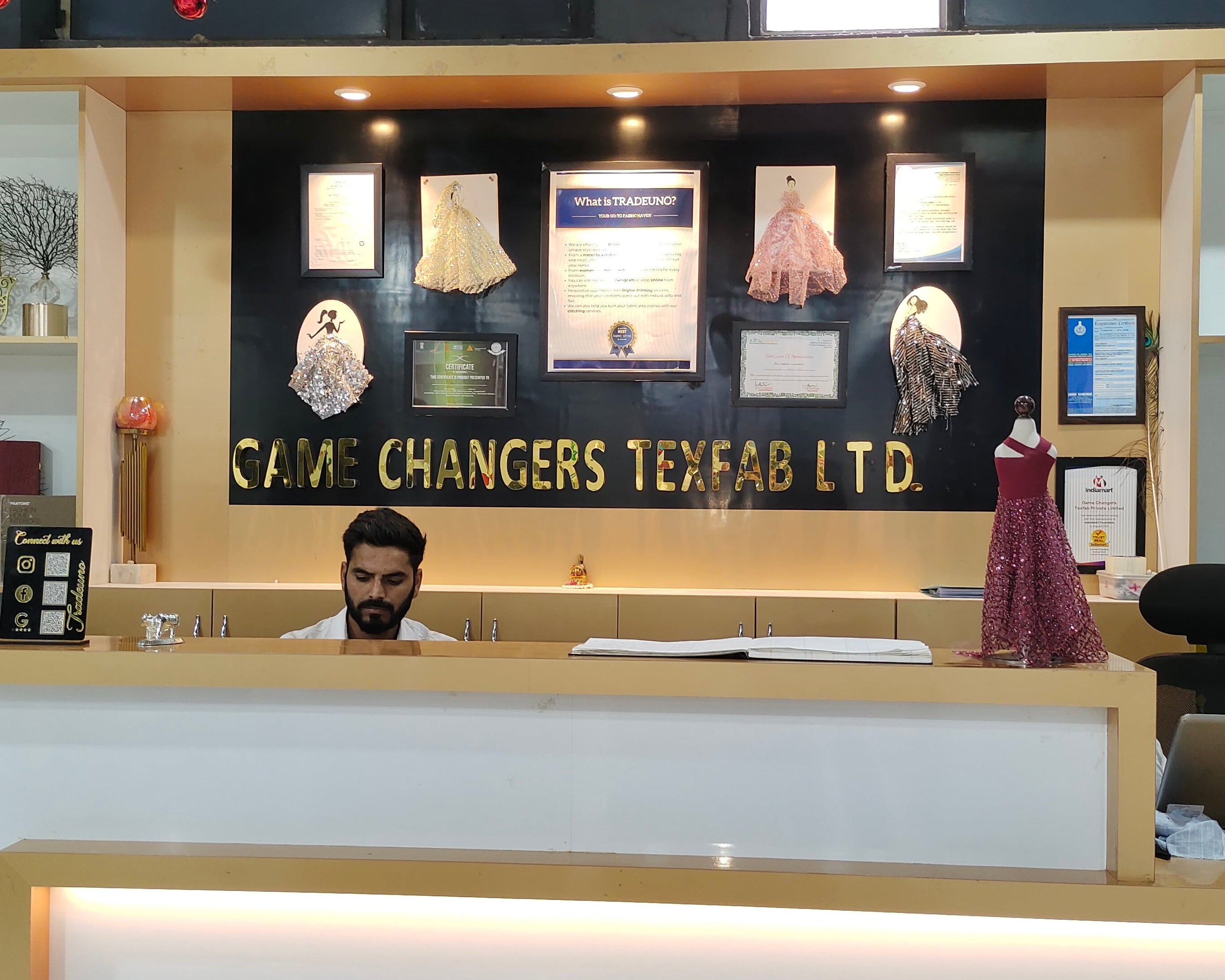 A modern textile factory reception desk with a man working behind it. The desk is white with light wood trim, and the back wall features a black panel with gold lettering that reads 'GAME CHANGERS TEXTILE LTD'.